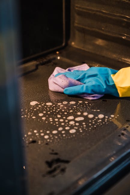 Close-up of a person wearing blue and yellow rubber gloves, cleaning the glass door of a black oven with a cloth. The oven's interior is partially visible, showing a reflective surface and bubbles of