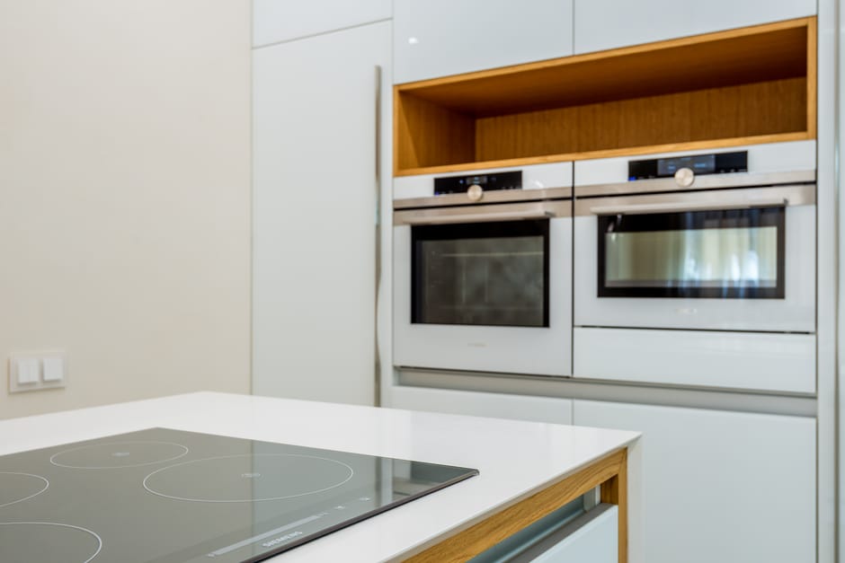 A modern kitchen with a white countertop and a built-in black glass induction cooktop, reflecting light for a clean appearance. In the background, there are two stainless steel wall ovens fitted into