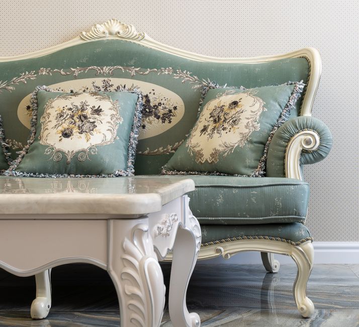A vintage-style sitting area featuring a light green upholstered sofa with ornate white wooden carvings and matching decorative pillows with floral embroidery, situated next to a white marble-topped c